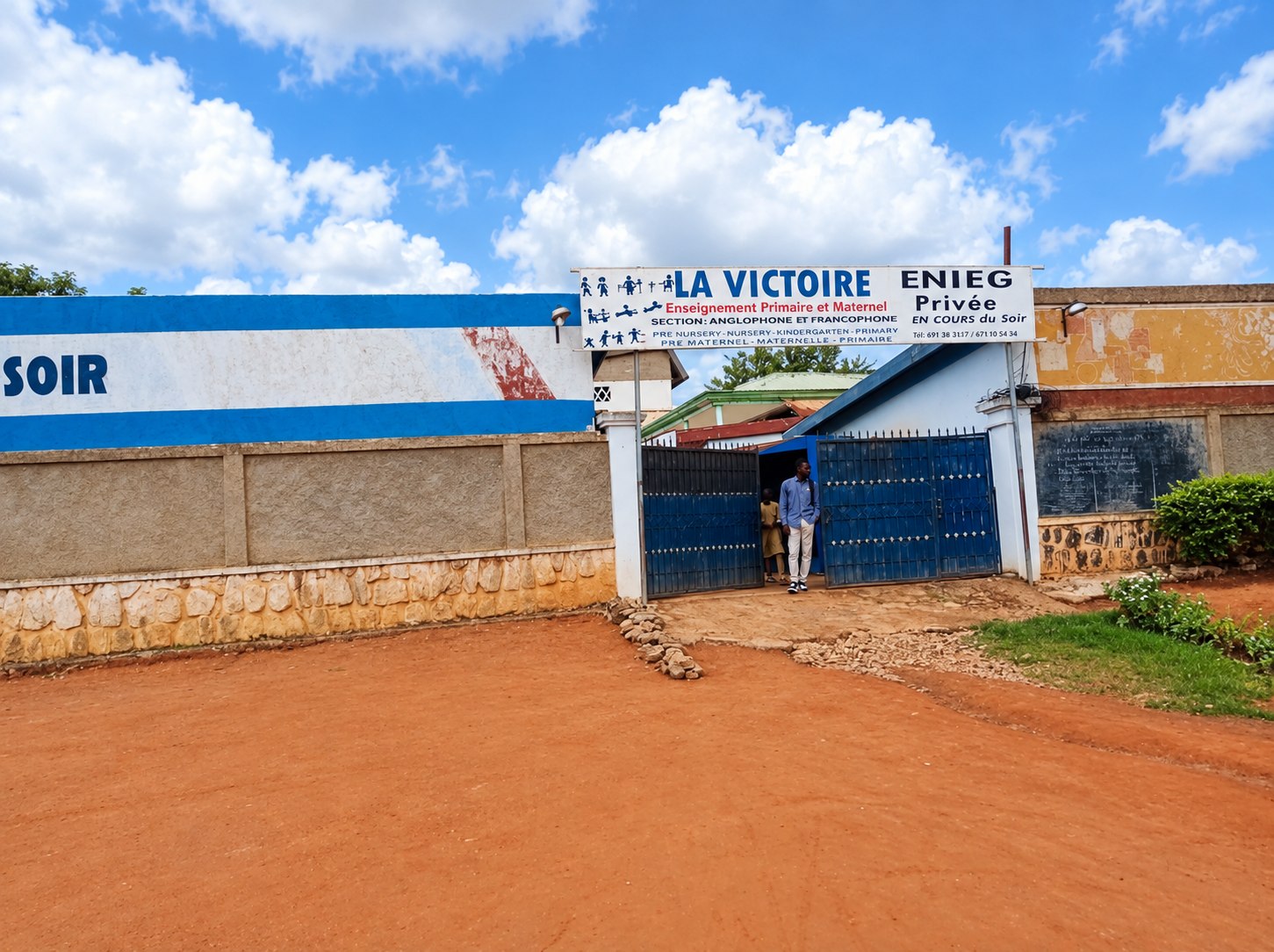 Entrée de LA VICTOIRE à Jolie Soir : enseigne « Enseignement primaire et maternel, section anglophone et francophone », portail bleu et sol de latérite.